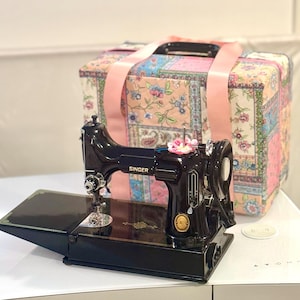 Puede incluir: Una máquina de coser Singer negra con una funda de transporte con estampado floral. La máquina de coser tiene una mesa abatible y un pedal.