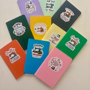 Puede incluir: Una colección de cuadernos coloridos con pegatinas con temática de costura. Los cuadernos son de varios colores, incluyendo verde, azul, rosa y amarillo. Las pegatinas presentan máquinas de coser, tijeras y frases como "Quilty Pleasure" y "Just Sew".