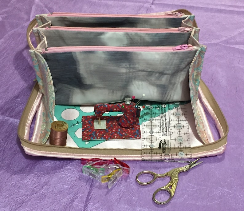 May include: A pink and gray fabric sewing kit with three zippered compartments, a small sewing machine, a spool of thread, a ruler, and a pair of scissors. The kit is open and the contents are visible.