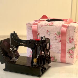 Peut inclure: Une machine à coudre Singer noire vintage avec des détails dorés et le logo Singer. Un sac de transport de machine à coudre à motifs floraux roses avec des poignées en ruban rose est derrière.