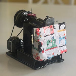 May include: Black sewing machine with a white fabric cover featuring a colourful pattern of sewing machines. The fabric cover has a floral design and the text "Sew Sweet" on it.