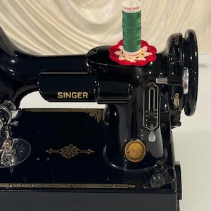 May include: A black Singer sewing machine with gold accents. The machine has a spool of green thread on top, and a red and white crocheted thread holder.