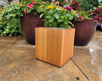 May include: A square wooden planter box with a light brown finish. The planter is made of vertical wood slats and has a green plastic liner inside. The planter is sitting on a brown tiled patio.