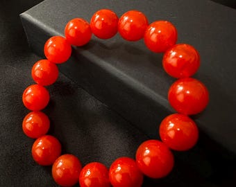 May include: A striking red beaded bracelet. The bracelet features large, round, glossy beads. The beads are strung together, forming a circular shape. The bracelet is presented on a black surface.