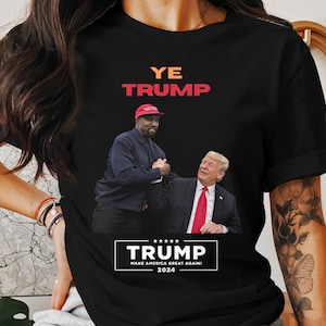 Puede incluir: Camiseta negra con un gr&aacute;fico de dos hombres d&aacute;ndose la mano, con el texto "YE TRUMP" en naranja y rojo. Debajo, una pancarta dice "TRUMP MAKE AMERICA GREAT AGAIN! 2024". La camiseta es de cuello redondo.