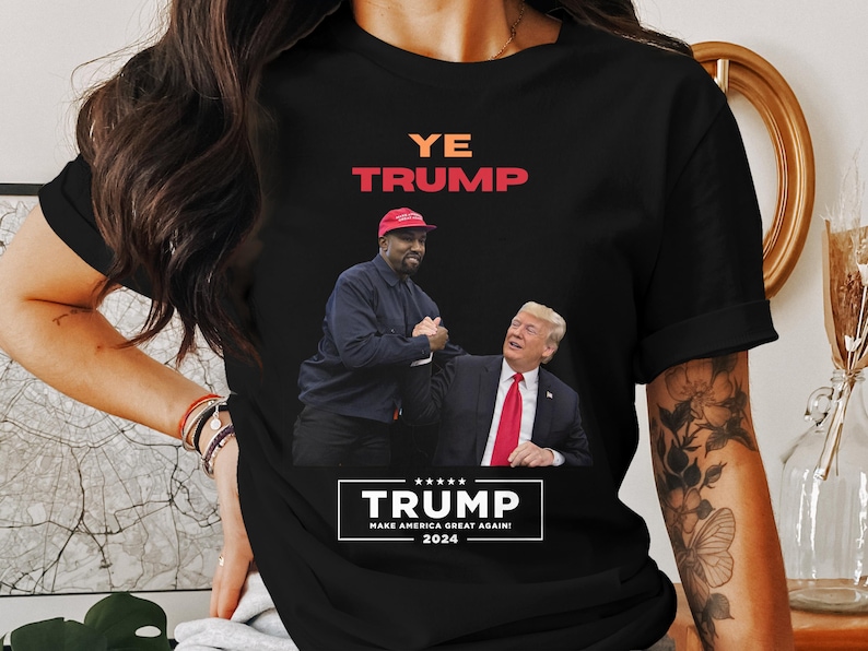 Puede incluir: Camiseta negra con un gr&aacute;fico de dos hombres d&aacute;ndose la mano, con el texto "YE TRUMP" en naranja y rojo. Debajo, una pancarta dice "TRUMP MAKE AMERICA GREAT AGAIN! 2024". La camiseta es de cuello redondo.