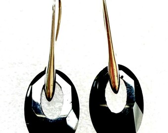 May include: A pair of black oval-shaped earrings with a faceted design. The earrings have a gold-colored hook and a polished finish, reflecting light. The jewelry is set against a white background.