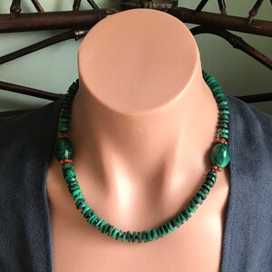 May include: A green malachite necklace with a dark green and black pattern. The necklace has two larger malachite beads and small red beads. The necklace is displayed on a mannequin with a blue top.