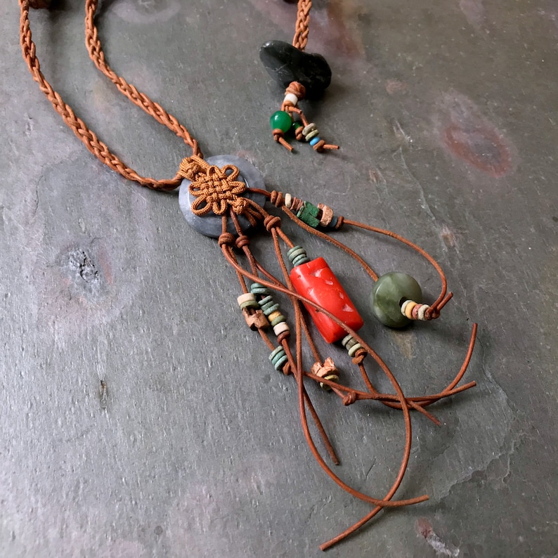 May include: A handcrafted necklace featuring a braided brown cord, a central gray stone with a decorative knot, and dangling leather strands adorned with red, green, and turquoise beads. A second necklace is visible in the background.