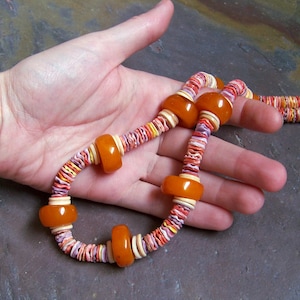 May include: A colorful necklace with orange, white, yellow, and purple accents. The necklace features large, round orange beads interspersed with smaller, multi-colored beads. The necklace is held in a hand.