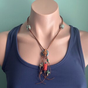 May include: A handcrafted necklace featuring a braided brown leather cord. The necklace has light blue square beads, a central blue pendant with a gold knot design, and a tassel of red, green, and blue beads.