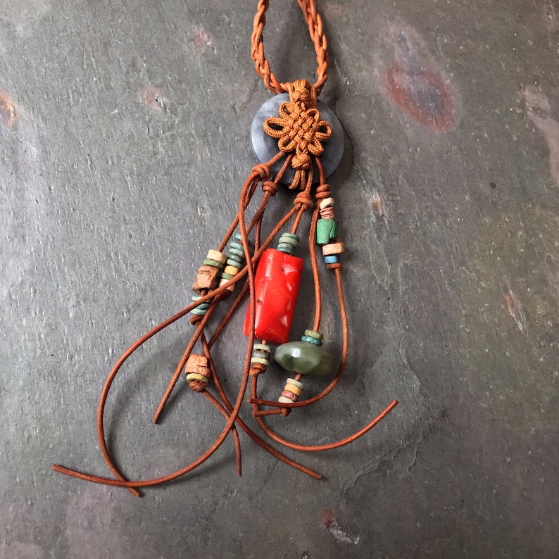 May include: A handcrafted necklace featuring a gray circular pendant with a brown woven knot. Brown leather cords cascade down, adorned with red, green, and turquoise beads. The necklace has a braided brown cord.