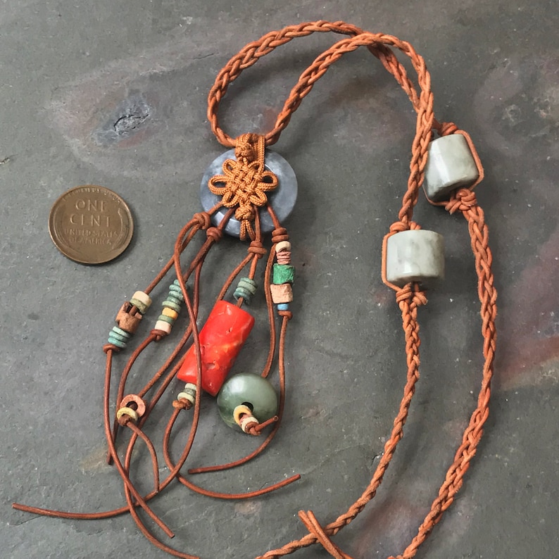 May include: A handcrafted necklace featuring a central blue stone with a decorative knot, adorned with leather strands and colorful beads. The necklace includes a red coral-like bead and a green stone bead. The necklace is displayed next to a US one-cent coin.