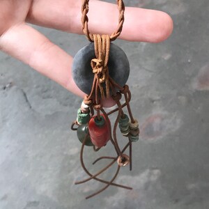 May include: A handcrafted necklace featuring a dark gray circular pendant with a braided brown leather cord. The pendant is adorned with brown leather strands and colorful beads in red, green, and cream colors. The necklace is held by a hand.