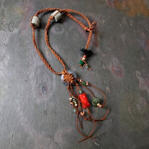 May include: A handcrafted necklace featuring a braided brown cord with gray stone beads. The necklace has a central pendant with a knot design and dangling strands adorned with red, green, and beige beads. The necklace is displayed on a gray surface.