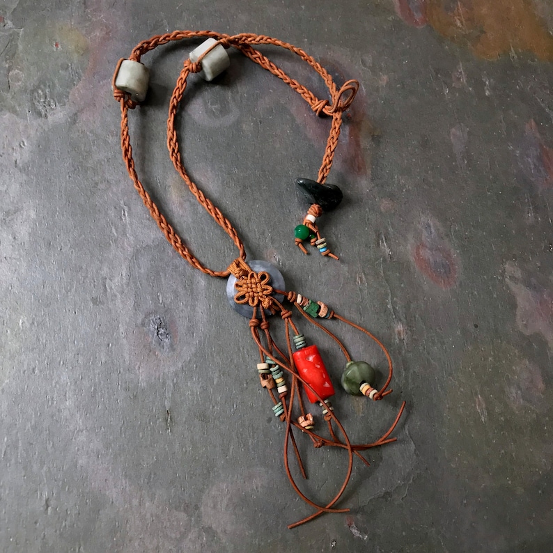 May include: A handcrafted necklace featuring a braided brown cord with gray stone beads. The necklace has a central pendant with a knot design and dangling strands adorned with red, green, and beige beads. The necklace is displayed on a gray surface.