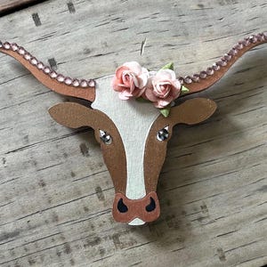 May include: Wooden cutout of a cow head with brown and white paint. The cow has pink flowers on its head and rhinestones on its horns.