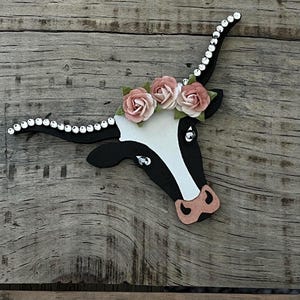 May include: A black and white wooden cutout of a cow's head with pink flowers on its head. The horns are decorated with rhinestones.