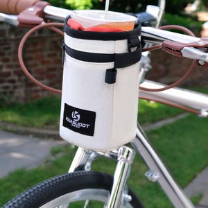 Puede incluir: Un portavasos blanco para manillar de bicicleta con cremallera y correa negras. El soporte tiene un parche negro con el texto "KUASUODT". Dentro hay un vaso de plástico transparente con pajita.