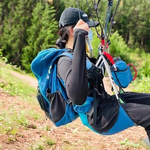 Puede incluir: Una persona con un arnés azul y negro y una mochila azul está conectada a un arnés de parapente. La persona lleva un sombrero negro y una mascarilla.
