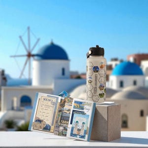 May include: A cream-coloured water bottle with a black lid and travel-themed graphics sits on a small, grey block. A travel journal and postcards are displayed nearby. The background shows white buildings and blue domes, reminiscent of a Greek island.