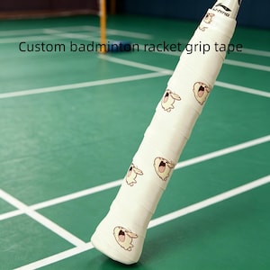 May include: A white badminton racket grip tape with a repeating cartoon character design. The grip tape has the text "Custom badminton racket grip tape" printed on it. The racket is on a green court.