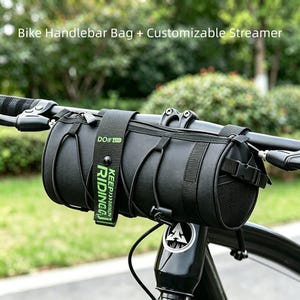 May include: Black handlebar bag attached to a bicycle. The bag is cylindrical with black straps and a buckle. A green tag with the words "KEEP RIDING" is attached. The text "Bike Handlebar Bag + Customizable Streamer" is at the top.