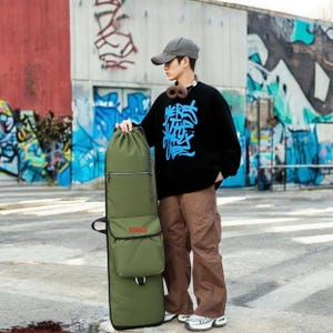 May include: An olive green snowboard bag with pockets and a drawstring. A person wears a black sweater with blue graffiti text, brown trousers, and a grey baseball cap. The bag is approximately 160 cm in length.