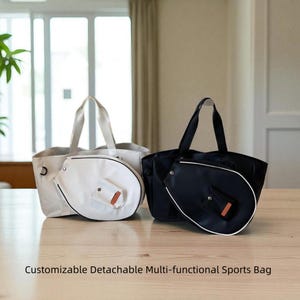 Puede incluir: Dos bolsas deportivas, una blanca y otra negra, se encuentran una al lado de la otra sobre una superficie de madera. Cada bolsa tiene un soporte desmontable para raqueta de tenis con un acento marrón. Las bolsas tienen asas.