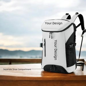 May include: A white and black backpack with the text "Your Design" printed on the front. The backpack has a separate shoe compartment and black zippers. The bag is sitting on a wooden surface.