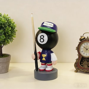 May include: A whimsical figurine of an 8-ball character wearing a blue and white uniform, a green cap with "Snooker Masters" text, and holding a pool cue. The figure stands on a gray base, with a gold trophy on the chest.