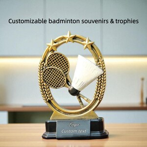 Op de afbeelding: Een goudkleurige badminton trofee met een zwarte basis. De trofee heeft een shuttlecock en twee rackets in een lauwerkransontwerp, bekroond met drie gouden sterren. De basis heeft ruimte voor een logo en aangepaste tekst. Tekst op de afbeelding: Aanpasbare badminton souvenirs & trofeeën.