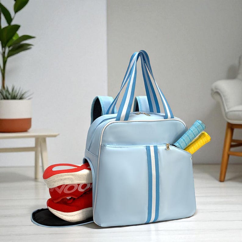 May include: A light blue sports bag with white and blue striped detailing. The bag features a side pocket containing two tennis rackets and a separate compartment for red and white athletic shoes. It has a top handle and shoulder straps.