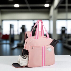 May include: A pink sports bag with white and pink striped handles and vertical stripes. The bag has a separate compartment for shoes and a pocket with two black tennis rackets. A white gym is visible in the background.