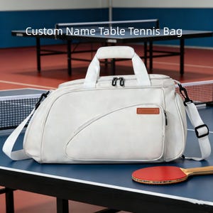 May include: A white table tennis bag with the text "Custom Name Table Tennis Bag". The bag has a top handle, a shoulder strap, and a small front pocket. A red and black ping pong paddle rests on the table.