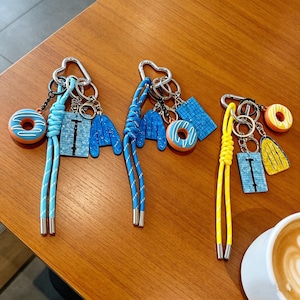 May include: Keychains with various charms, including a blue donut with white icing, a blue jacket, and a blue square. The keychains have silver hardware and colorful cords. One keychain has a yellow cord.