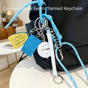 May include: A custom name swim themed keychain attached to a black backpack. It features a yellow swim fin charm, a blue grid pattern charm, a white retractable measuring tape, and a black cord with white letter beads spelling out SUNDAY.