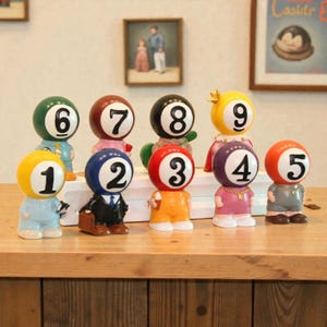 May include: A collection of whimsical billiard ball figurines, each with a different number from 1 to 9. The figurines are dressed in various outfits and colors, including blue, black, red, and yellow. They are arranged on a wooden surface.