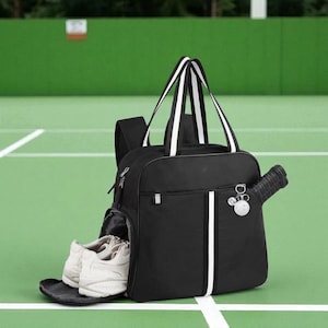May include: Black tennis bag with white stripes and handles. A tennis racket is in the side pocket, and a pair of white tennis shoes are in the front pocket. The bag has a small charm and is on a green tennis court.