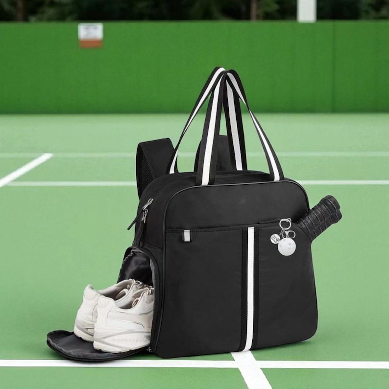 May include: Black tennis bag with white stripes and handles. A tennis racket is in the side pocket, and a pair of white tennis shoes are in the front pocket. The bag has a small charm and is on a green tennis court.