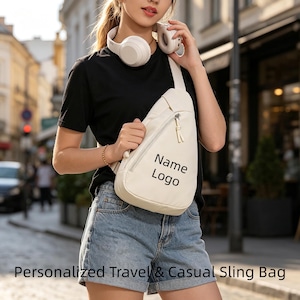 May include: A cream-coloured sling bag with a single shoulder strap and a diagonal zippered pocket. The front of the bag features the black text Name Logo. The bag is worn across the chest, constructed from a smooth material with a matching zipper pull.