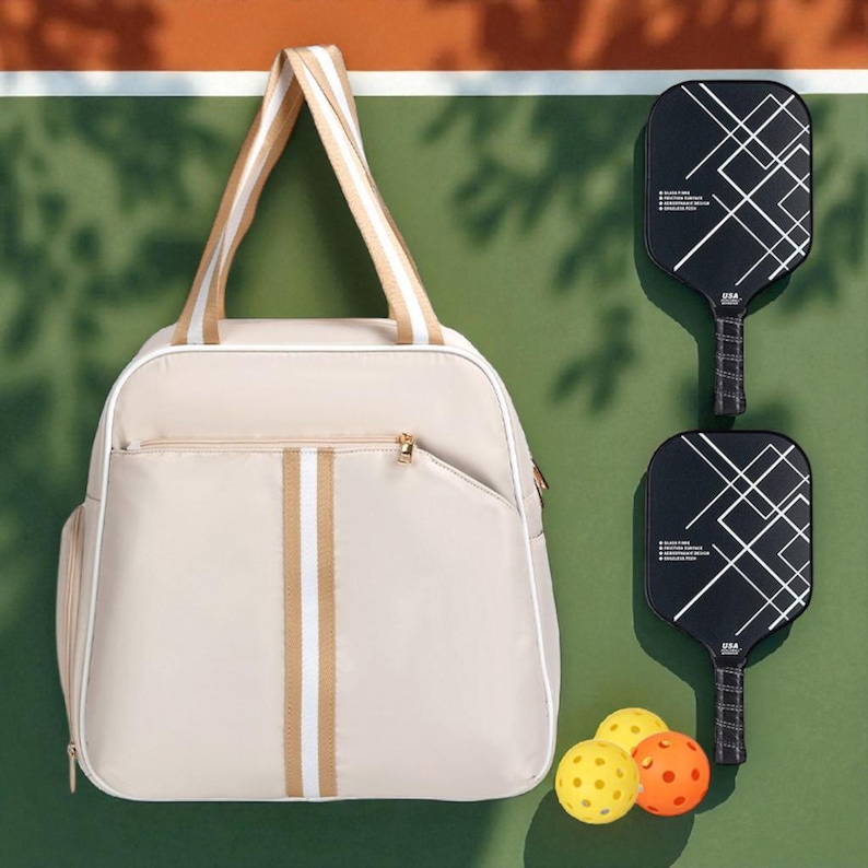 May include: A beige pickleball bag with white and tan stripes, two black pickleball paddles with white geometric designs, and three pickleballs. The bag has a zippered pocket and a tan and white striped handle. The paddles have black grips.