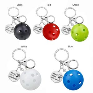 May include: A collection of pickleball keyrings in various colours: black, red, green, white, and blue. Each keyring features a small pickleball and a charm with the text "PEACE LOVE PICKLEBALL". The keyrings have a silver keyring.