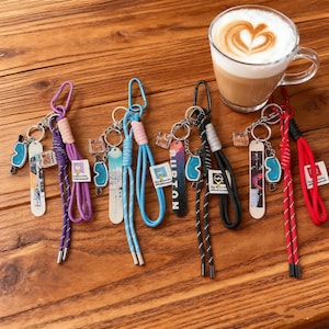 Puede incluir: Una colección de llaveros con colgantes de mini snowboard en varios colores, incluyendo morado, azul y rojo. Cada llavero tiene un cordón a juego y un cierre metálico. También hay una taza de café con arte latte.