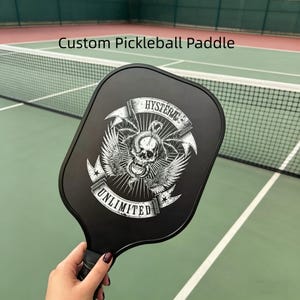 May include: Black pickleball paddle with a skull and wings graphic, the text "HYSTERIC UNLIMITED" is displayed on the paddle. The paddle is held in front of a pickleball court. The text "Custom Pickleball Paddle" is displayed above the paddle.