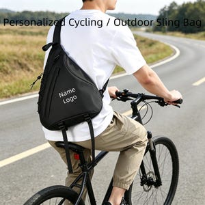 May include: A black triangular sling bag with a single shoulder strap, featuring the text Name Logo printed on the front. The bag includes a vertical zippered pocket, an adjustable strap system, and black elastic cord detailing on the side.