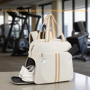 May include: Cream pickleball paddle bag with gold stripes and handles. The bag features a front zipped pocket and a side pocket holding a black paddle. White trainers are visible in a side compartment. Includes a customizable name pendant.