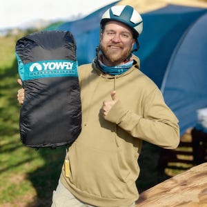 Può includere: Una sacca da parapendio nera e turchese con la scritta "YOWFLY PARAGLIDING" è tenuta da una persona che indossa una felpa con cappuccio beige e un casco bianco e turchese. La persona sorride e fa il pollice in su.