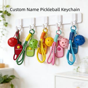May include: A display of vibrant pickleball keyrings suspended from a white wall fixture. Each keyring showcases a pickleball, a tag, and a loop. The keyrings are in red, green, yellow, pink, and blue. The text "Custom Name Pickleball Keychain" is visible above.