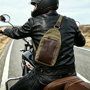 May include: A crossbody sling bag featuring a khaki canvas body with a dark brown leather front pocket and a matching adjustable shoulder strap. The bag has a zippered closure and is worn over a black leather jacket while riding a motorcycle.